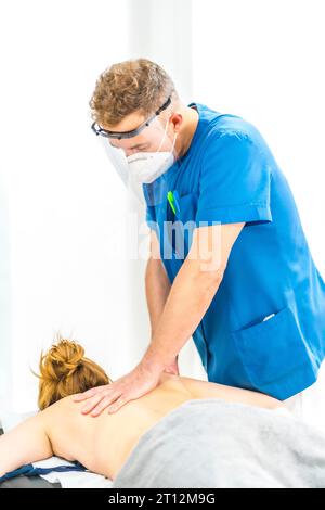 Physiotherapist with mask and screen performing a massage with two hands on the back of a girl. Physiotherapy safety measures in the Covid-19 Stock Photo