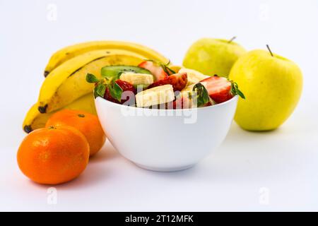 A fruit salad cut in a white bowl on a white background next to tangerines, bananas and apples Stock Photo