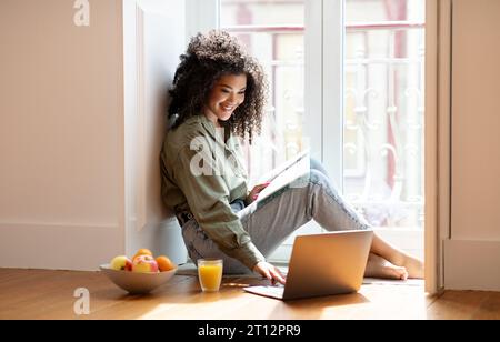 Young black lady using laptop holding notebook for notes indoor Stock Photo