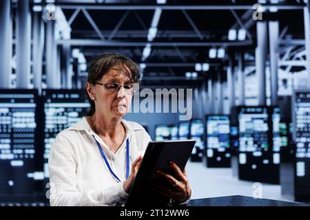 Senescent developer updating data center equipment, writing intricate binary code scripts on tablet, using programming to fortify cybersecurity in technically advanced server hub Stock Photo