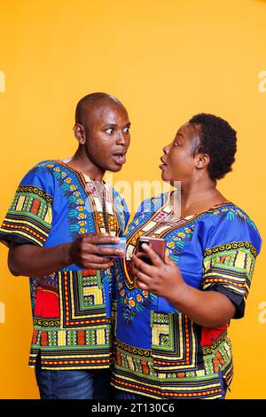 Shocked couple looking at each other with eyes wide open while checking price in online store using smartphone. Surprised african american man and woman shopping with mobile phone Stock Photo