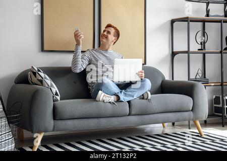 Young man with laptop switching on air conditioner at home Stock Photo