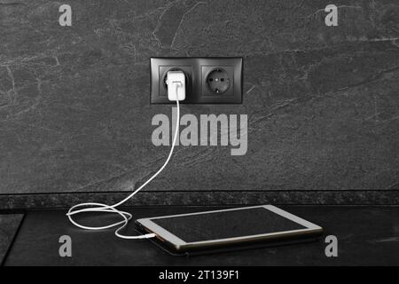 Tablet charging from electric socket on dark grey table indoors Stock Photo