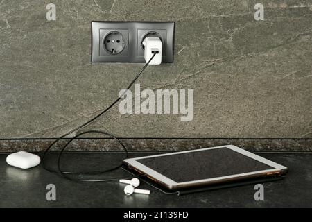 Tablet charging from electric socket on dark grey table indoors Stock Photo