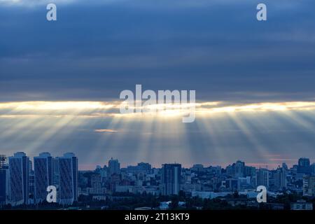 The sun's rays at dawn in the blue break through the thick clouds over the sleeping city. Shining light in a dramatic morning sky. Stock Photo