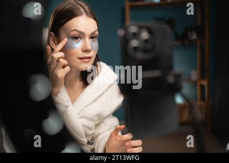 Cosmetologist vlogger recording a video. Blogger putting under eye masks for puffiness, wrinkles, dark circles. Eye patches concept. Satisfied woman a Stock Photo