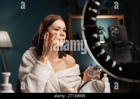 Young influencer woman creating social media videos while applying cream face mask recording video on smartphone. Technology, cosmetics and fashion co Stock Photo