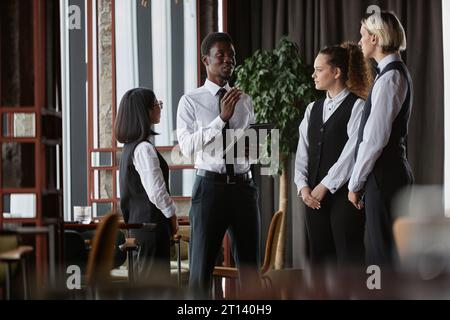 Staff meeting in luxury restaurant with manager talking to three servers wearing classic uniform in dining room Stock Photo