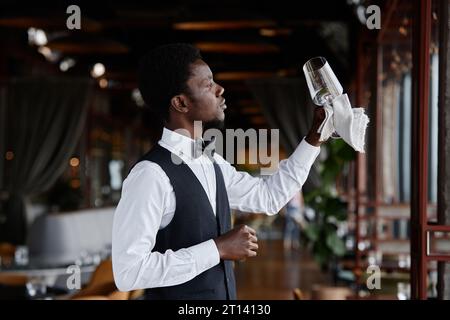 Side view portrait of Black young man as elegant waiter holding crystal glass in restaurant interior, copy space Stock Photo