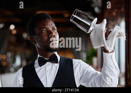 Portrait of Black young man as elegant waiter polishing glasses in luxury restaurant, copy space Stock Photo