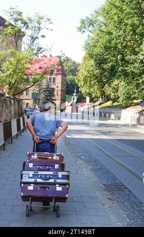 The man pulls old suitcases on a wheelbarrow . Elderly man pulls a wheelbarrow with suitcases. Heap Of Old Suitcases, Trolley Suitcases. The concept o Stock Photo