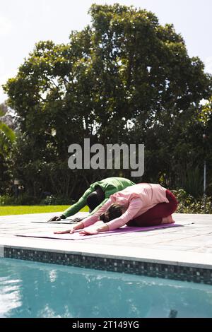 Diverse couple practicing yoga stretching next to swimming pool in sunny garden, copy space Stock Photo