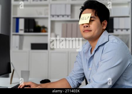 Young adult Asian businessman having HELP sticky note on his face in the office setting background. Business man is bored, frustrated, unhappy, and workaholic. Stock Photo