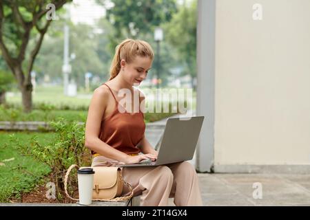 Smiling young woman sitting on bench in park and working on laptop Stock Photo