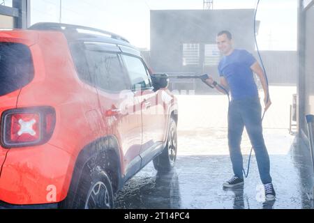 Young man washes his car with high pressure water outdoors in a car wash Stock Photo