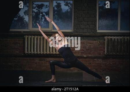 Beautiful sporty fit yogini woman practices yoga asana Utthita Parshvakonasana in the dark hall. Stock Photo