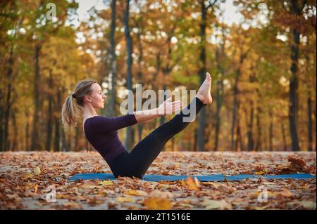 Beautiful young woman practices yoga asana Paripurna Navasana Pose on the wooden deck in the autumn park Stock Photo