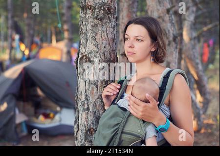 A young mother in the forest with her baby in a sling. Camping on a background. Stock Photo