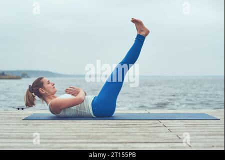 Beautiful young woman practices yoga asana Paripurna Navasana on the wooden deck near the lake. Stock Photo