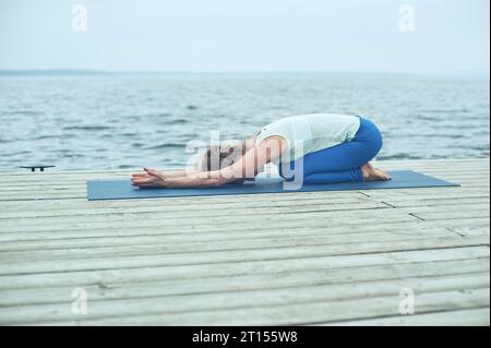 Beautiful young woman practices yoga asana Gomukhasana - Cow face pose on the wooden deck near the lake. Stock Photo