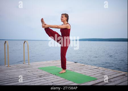 Beautiful young woman practices yoga asana Utthita Hasta Padangushthasana on the wooden deck near the lake. Stock Photo