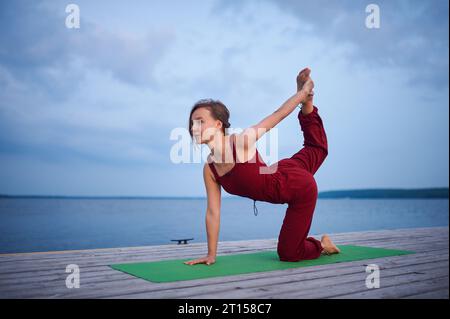 Beautiful young woman practices yoga on the wooden deck near the lake. Stock Photo