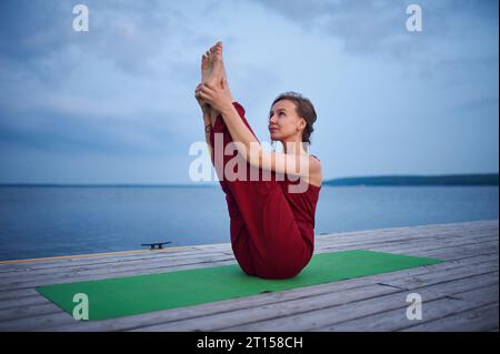 Beautiful young woman practices yoga asana Paripurna Navasana Pose on the wooden deck near the lake. Stock Photo