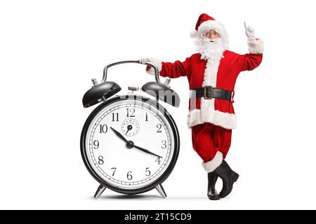 Santa claus leaning on a big alarm clock and pointing up isolated on white background Stock Photo