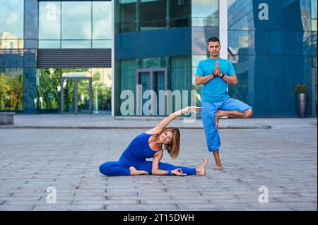 An attractive young woman and man doing yoga outdoors against the background of a modern city. Stock Photo