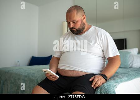 Fat bearded bald guy with smartphone chatting, dials number, types message, watches news, weather Stock Photo