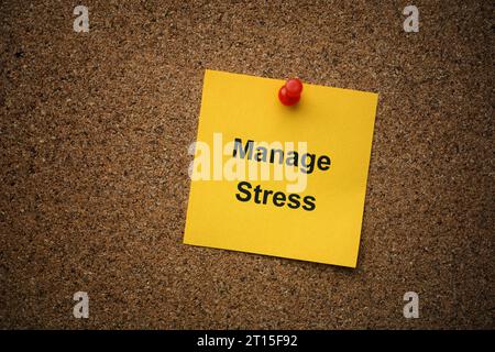 A yellow paper note with the words Manage Stress on it pinned to a cork board. Close up. Stock Photo