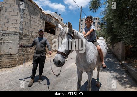 Bayt Surik, Palestine – June 19, 2023: Happy Young Arab Boy Rides a Beautiful White Horse and Smiles While a Man Holds the Strap in a Small Street Stock Photo