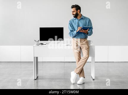 Confident indian businessman sitting leaning on table and looking at computer with blank screen in office, free space Stock Photo