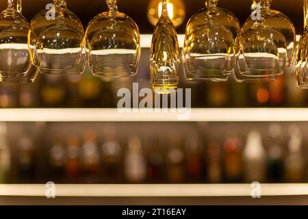 Clean wine glasses hanging over the bar counter on out of focus background with shelves with bottles of alcoholic beverages. Stock Photo