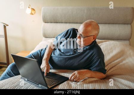 An elderly man is lying on his bed, having fun with his laptop, working remotely, surfing social networks on the Internet. Stock Photo