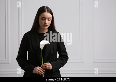 Sad woman with calla lily flower near white wall, space for text. Funeral ceremony Stock Photo