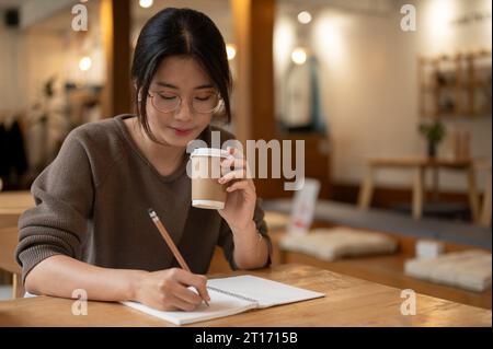 A pretty Asian woman focuses on keeping her diary or making lists in a book and sipping coffee while sitting at a table in a beautiful coffee shop. Stock Photo
