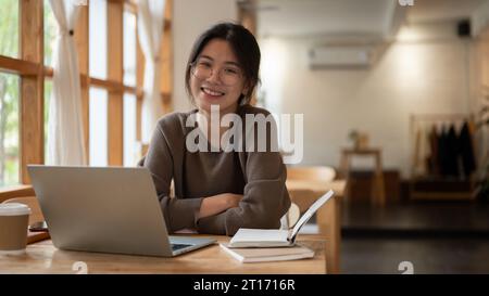 A portrait of a charming and smiling young Asian woman wearing eyeglasses sitting at a table with her laptop and books in a minimal coffee shop. Stock Photo