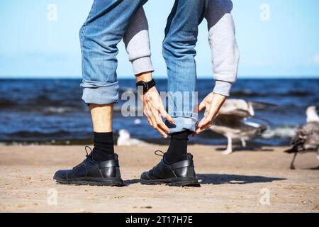 On the seashore of blue color stands a man in denim pants with black shoes in his hands Stock Photo