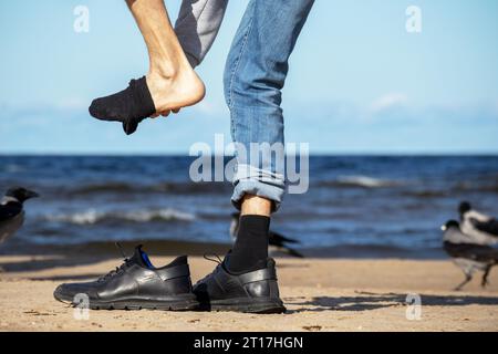 On the seashore of blue color stands a man in denim pants with black shoes in his hands Stock Photo