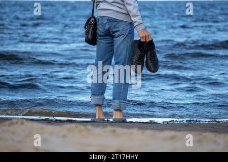 On the seashore of blue color stands a man in denim pants with black shoes in his hands Stock Photo