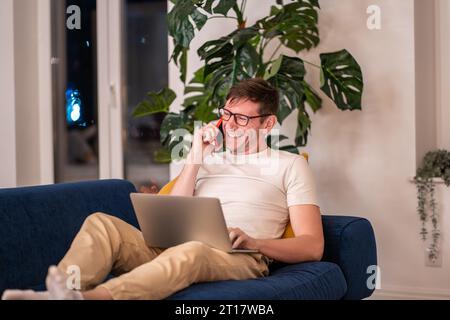 Laughing man in eyeglasses sitting on sofa with laptop on knees having fun cellphone conversation Stock Photo
