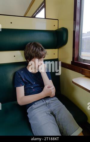 Child sitting alone and looking out the window of the old train car, boy has fist on chin and mouth, serious staring face thinking concept Stock Photo