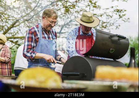 Two cheerful seniors share a playful moment while grilling at a barbecue during an outdoor picnic. Wearing aprons, they enjoy their time, with friends Stock Photo