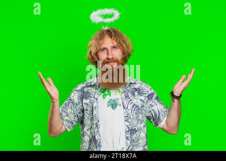 Portrait of smiling shy angelic young caucasian man with angel halo nimb over head flirting, looking at camera, positive love emotions celebrating holiday. Redhead bearded guy on chroma key background Stock Photo