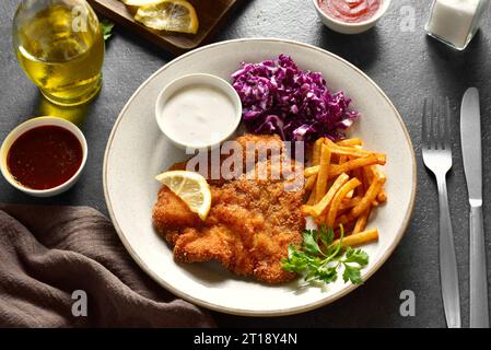 Schnitzel with potato fries, red cabbage salad and sauce on white plate over dark stone background. Close up view Stock Photo
