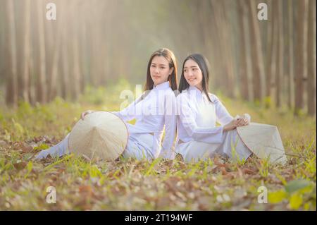 Two women in in traditional Vietnamese tunic dresses (ao dai) and hats (non la) sitting back to back in a forest, Vietnam Stock Photo