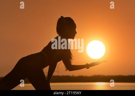 Silhouette of a woman listening to music while doing yoga by a lake at sunset, Thailand Stock Photo
