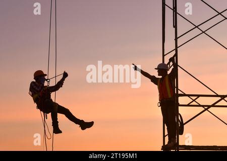 Silhouette of two construction workers on scaffolding and hanging on a harness on a construction site, Thailand Stock Photo