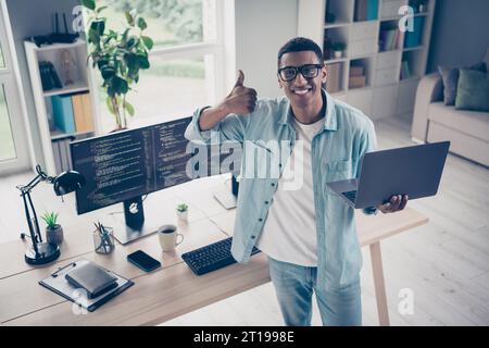 Photo of junior it specialist young guy web products developer holding laptop thumb up gesture enjoy his programming job in office Stock Photo